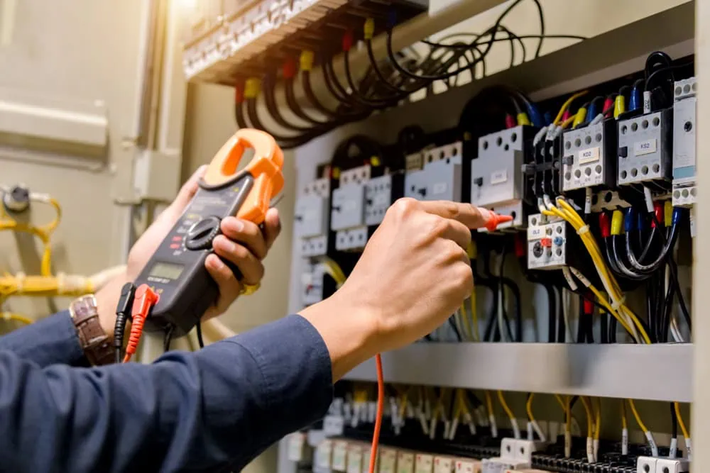 A technician's hands are shown using an electrical multimeter to test a circuit in an industrial control panel. The panel is filled with numerous wires and components. The Electrician Taylor County is holding the multimeter in one hand and using probes to check connections with the other.