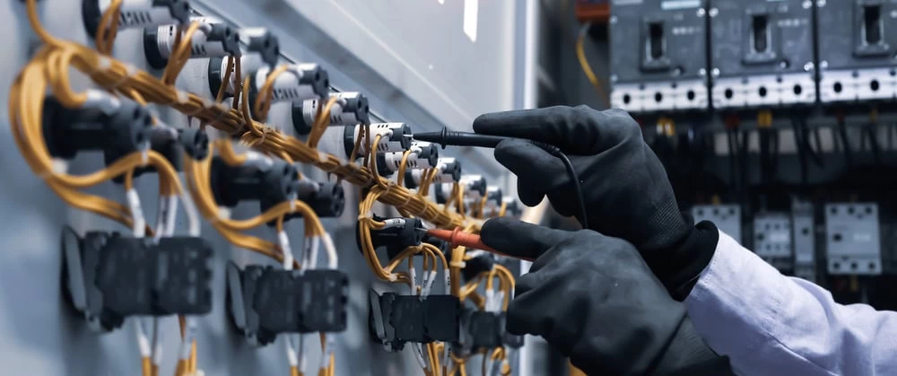 Close-up of an *Electrician in Taylor County* wearing gloves, working on electrical components with various cables and connectors on a panel. The individual is using a tool on one of the connectors, indicating detailed and precise electrical or technical work.