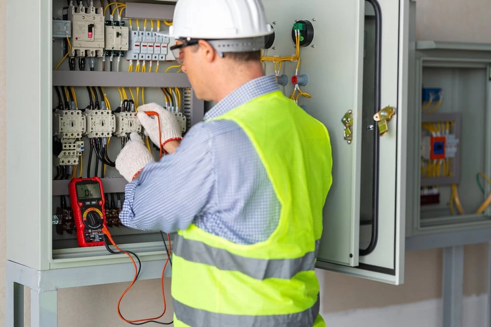 An Electrician in a neon yellow safety vest, blue shirt, safety gloves, and a white hard hat is using a multimeter to inspect electrical components inside an open control panel. Various wires and circuit breakers are visible inside the panel. This professional represents Electrical Contractor Taylor County's commitment to safety and precision.
