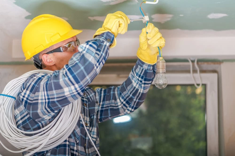 A worker in a yellow hard hat and gloves installs a ceiling light fixture. He wears protective eyewear and a checkered shirt, carrying a coiled electrical wire over his shoulder. Through the window with greenery outside, you can almost feel the expertise of an Electrician Taylor County prides itself on.
