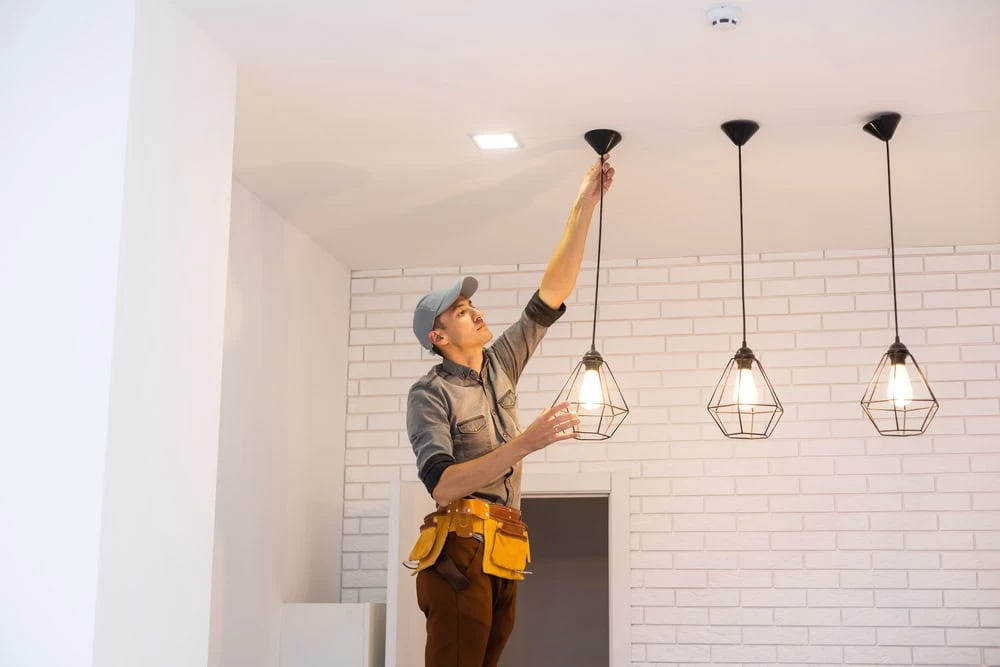 A person wearing a gray cap and a tool belt is standing on a ladder, adjusting one of three hanging light fixtures in a white room with a brick accent wall. The light fixtures, installed by an electrical contractor in Taylor County, have a geometric wireframe design and are illuminated.
