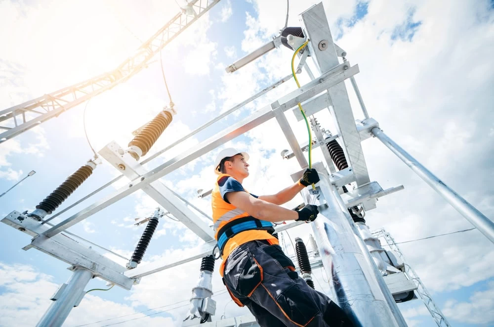 A utility worker, wearing a safety helmet, gloves, and an orange safety vest, is elevated on a platform as he inspects or repairs electrical equipment on a utility pole. The sky is clear with some clouds, and the sunlight is bright. Electrician Taylor County ensures all tasks are completed safely.