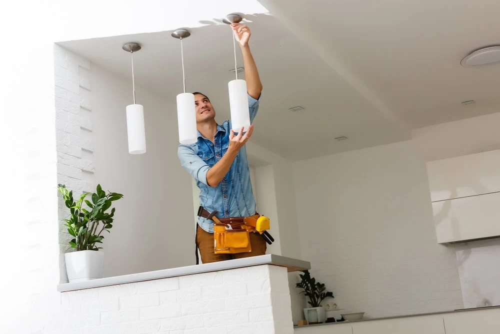 A person wearing a denim shirt and a tool belt, likely an electrician from Taylor County, is installing or adjusting a hanging light fixture in a modern, white kitchen. There are potted plants on a shelf against a white brick wall in the background.