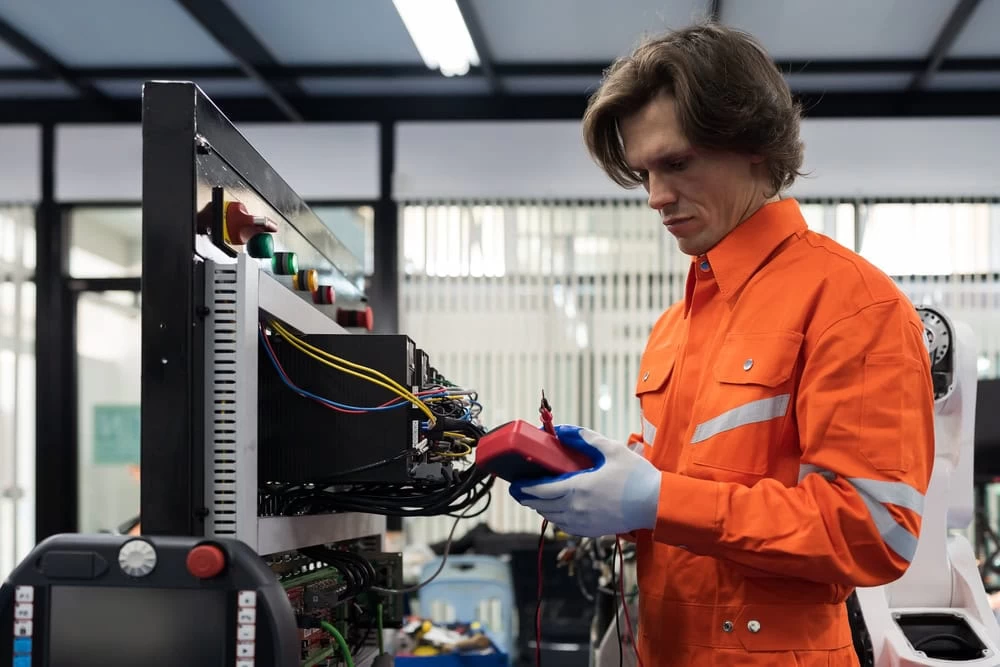 Electrical Technician Testing Taylor County Tx a Person with Brown Hair Wearing an Orange Work Jumpsuit and Gloves Uses a Multimeter to Test Wiring in an Electrical Panel in an Industrial Setting Various Wires and Switches Are Visible with Blurred Backgrounds of Machinery and Windows Showcasing the Expertise of an Electrical Contractor Taylor County