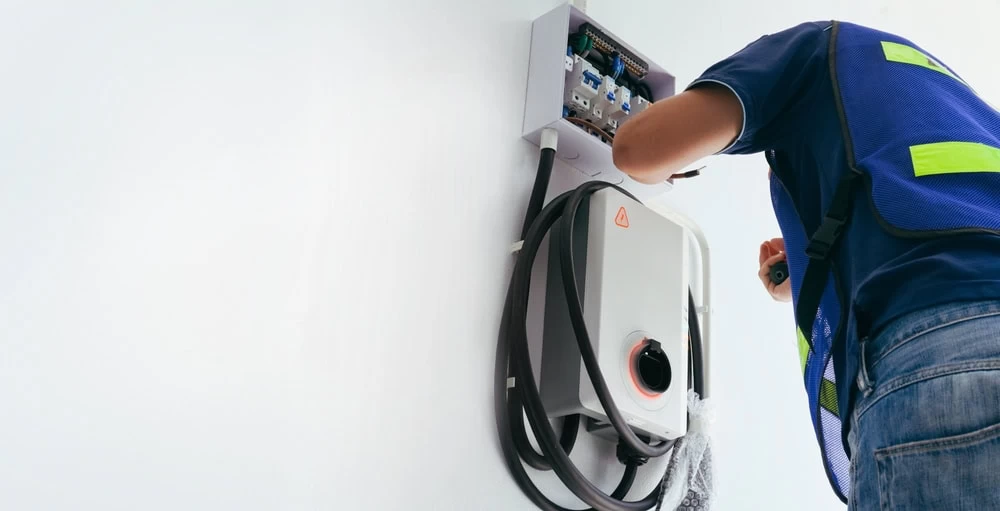 A person in a blue safety vest is meticulously installing or repairing an EV charger on the wall. The device, with its cables and control panel, demands their full attention as they focus intently on the task with their back partially turned.