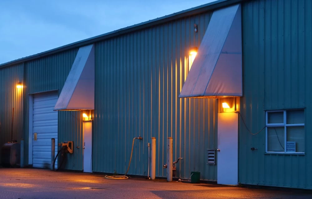 A teal industrial building with security lighting features two lit awnings above doors at dusk. The exterior is bathed in yellow light, highlighting a large garage door on the left, with hoses and utility connections nearby. A small window is nestled on the far right.