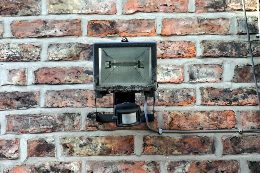 A rectangular security lighting fixture with a motion sensor is mounted on a textured brick wall. The weathered and slightly dusty light is positioned centrally on the multicolored bricks, enhancing the outdoor safety with its vigilant presence.