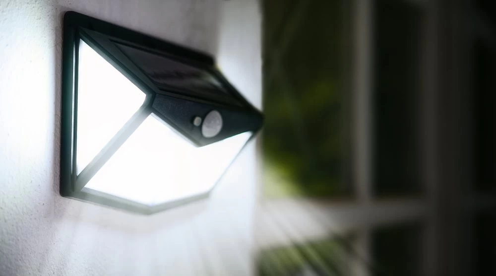 A wall-mounted, solar-powered LED light serves as essential security lighting, casting bright beams outward in the dark. The triangular design features a small sensor at its center and is angled toward a window with blurred surroundings in the background.