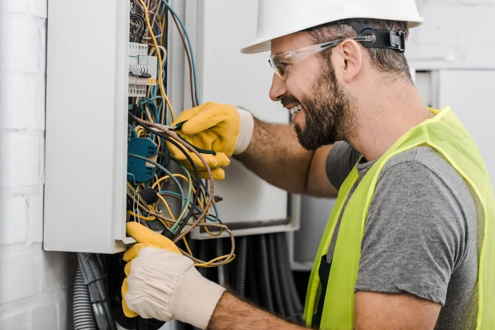 Man wearing a white hard hat, safety goggles, yellow vest, and gloves works on electrical wires in an open circuit breaker panel. He is smiling and using a tool to adjust the wires inside the panel.