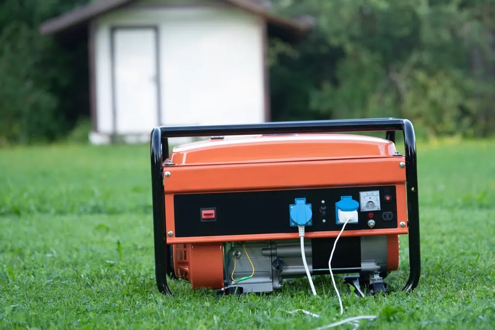 A portable orange electric generator sits on grass in the foreground, with two cords plugged into its outlets. A small structure with a white door and brown roof is blurred in the background, surrounded by greenery.