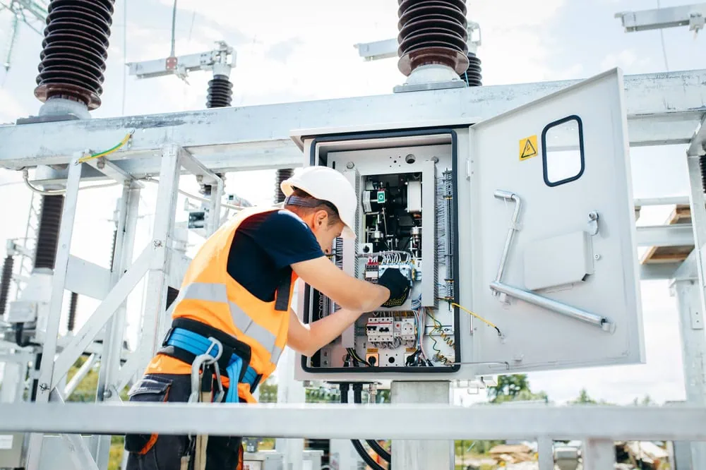 A worker in a hard hat and high-visibility vest is conducting electrical inspections, meticulously adjusting controls inside a metal panel at an outdoor power substation. The sky is clear, and various electrical components are visible in the background.