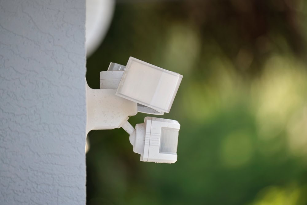 A white security floodlight, part of an advanced security lighting system, is mounted on the corner of a light gray wall. The dual lights and sensors are visible, set against a blurred green and brown outdoor background.