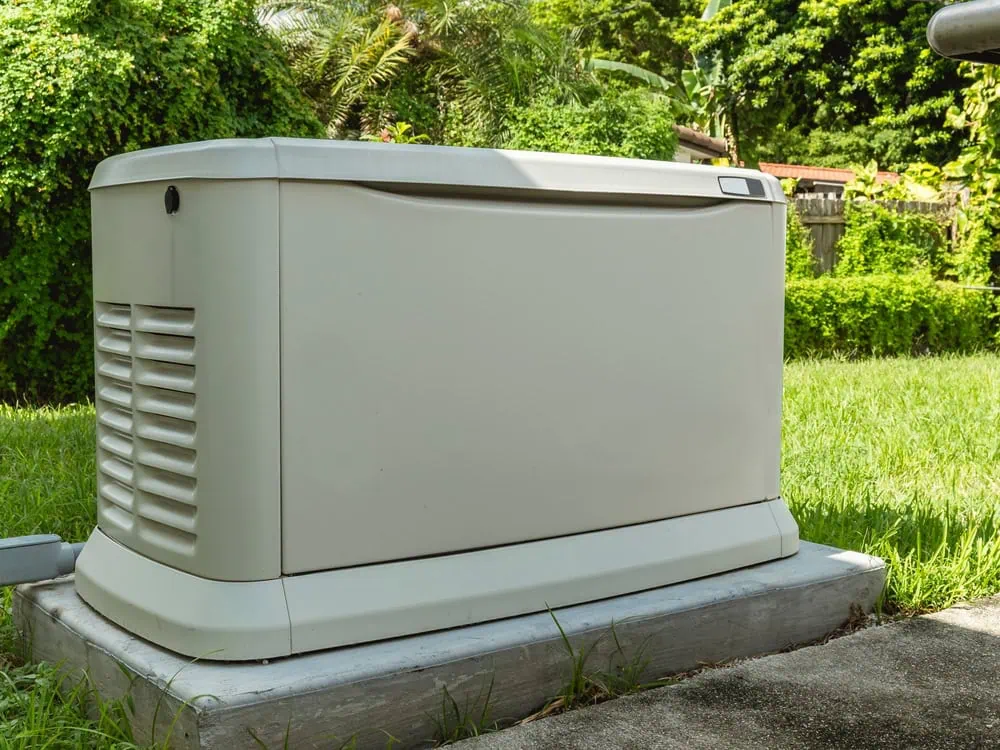 A beige generator, expertly installed on a concrete platform, sits in a grassy yard with dense greenery behind it. Its vented sides and sleek design allow it to blend seamlessly with the natural surroundings.