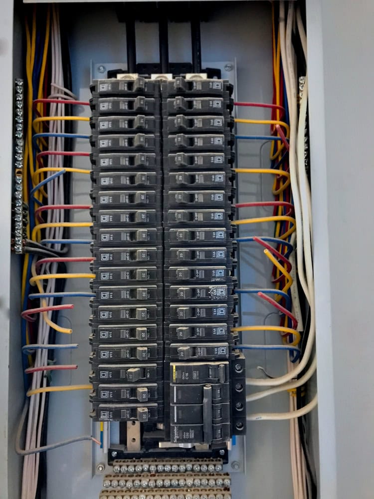An open electrical panel showcases multiple circuit breakers arranged vertically, highlighting recent circuit breaker panel upgrades. Each breaker is connected to neatly organized wires in red, yellow, white, and blue within the grey enclosure.