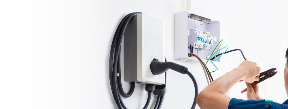 A person is performing an EV Charger installation on a white wall. They are using tools to connect wires to a circuit breaker, ensuring everything is securely in place. The charging cable is coiled neatly around the unit, ready for its first use.