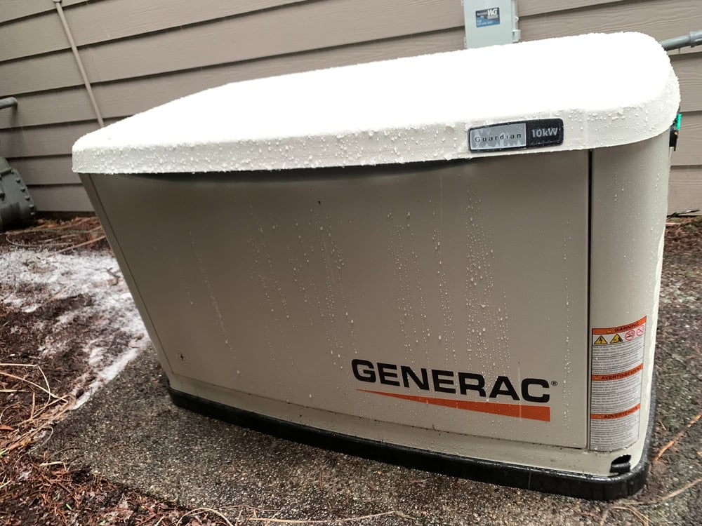 A Generac Guardian 16kW standby generator is expertly installed outside a building. The gray unit, covered with a light layer of snow and droplets of water, rests securely on a concrete pad beside a wall. This precise generator installation ensures reliable service even in wintry weather.