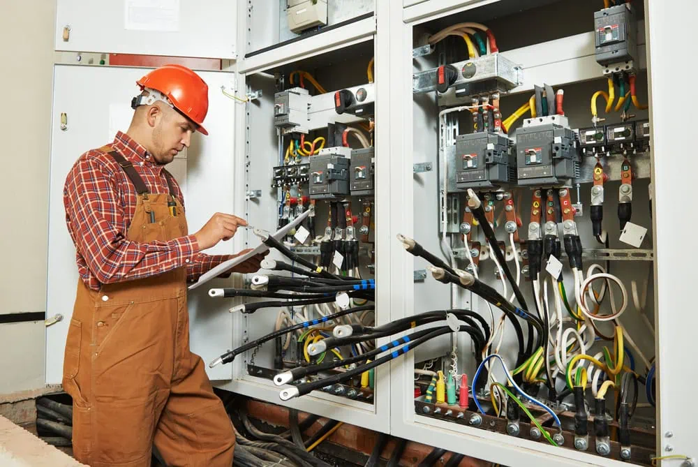 A worker in a red hard hat and brown overalls inspects electrical installations inside a circuit board cabinet. He holds a clipboard and pen, surrounded by various cables and components in the technical room.