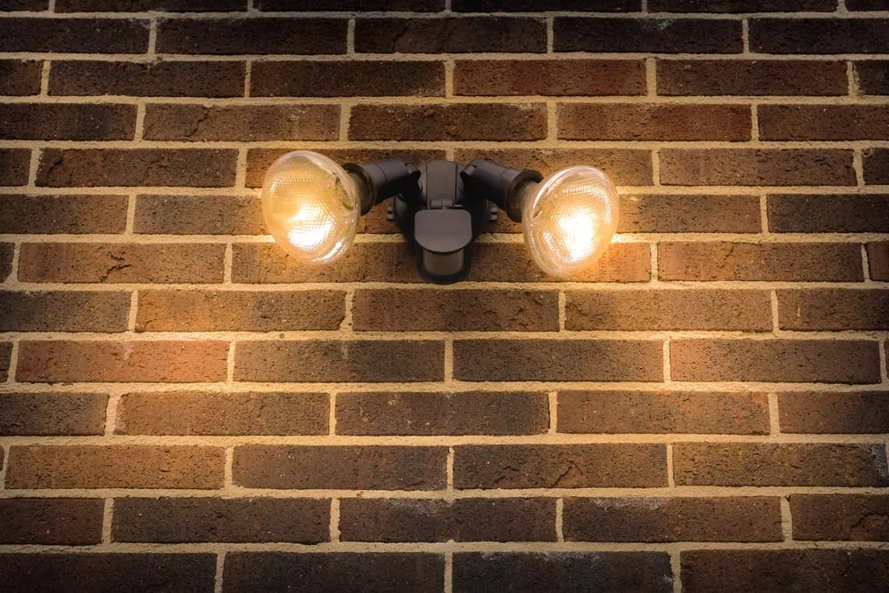 A dual-bulb outdoor light fixture serves as security lighting, mounted on a brown brick wall. Both bulbs are illuminated, casting a warm glow on the bricks, highlighting their texture and color.