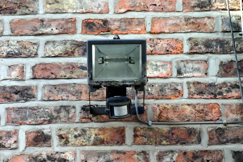 A rectangular security lighting fixture with a motion sensor is mounted on a textured brick wall. The weathered and slightly dusty light is positioned centrally on the multicolored bricks, enhancing the outdoor safety with its vigilant presence.