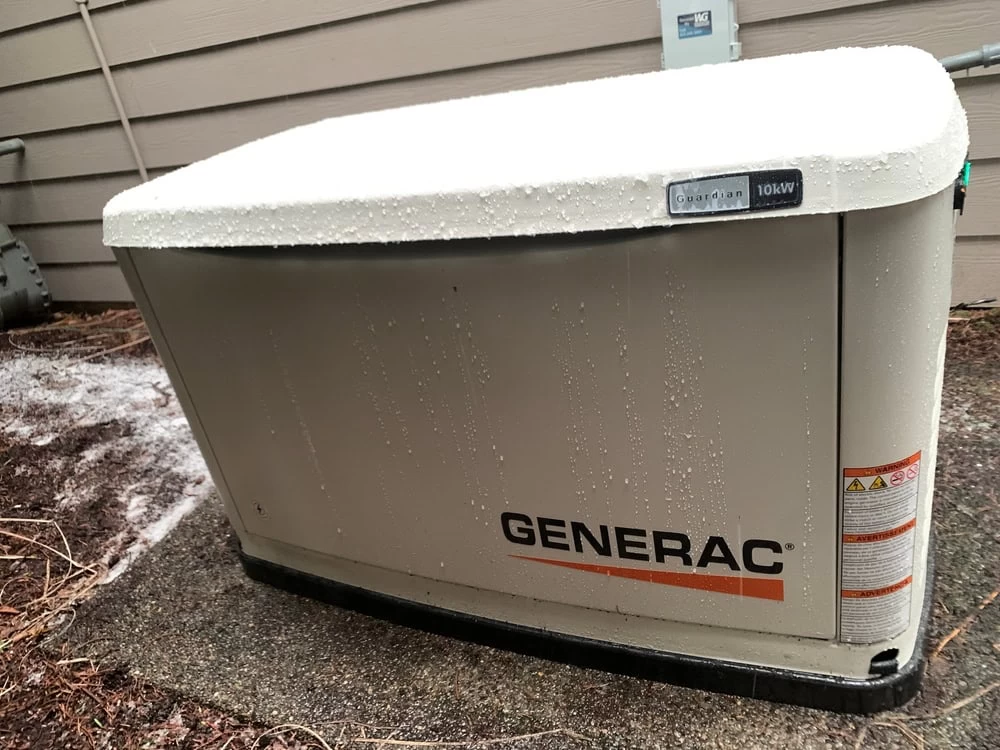 A Generac generator installation is visible outside a building, dusted lightly with snow. The beige unit is marked "Guardian 10kW" and "Generac" in black with an orange line underneath. A wooden siding serves as the backdrop.
