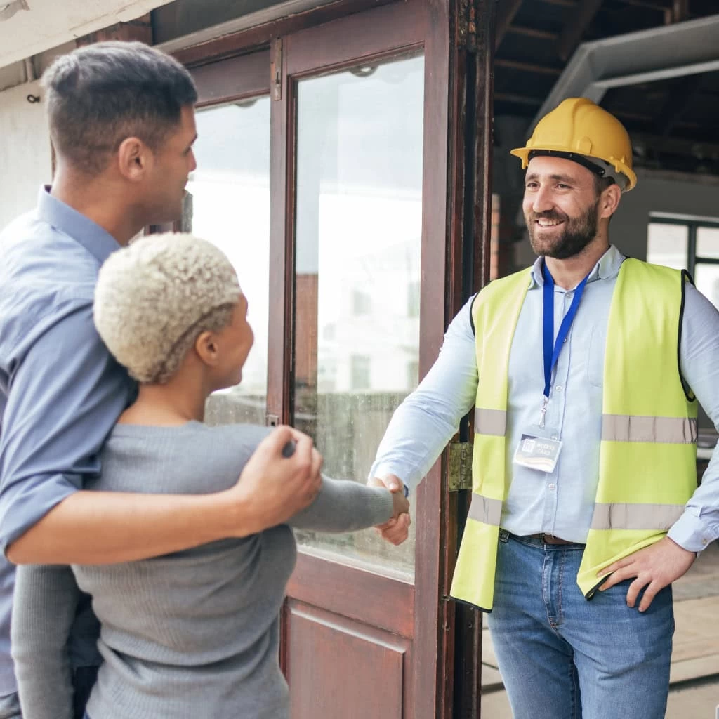 A construction worker wearing a yellow hard hat, yellow safety vest, and ID badge smiles while shaking hands with a couple. The man and woman in the couple are standing side by side, with the man’s arm around the woman. They are outside a building.
