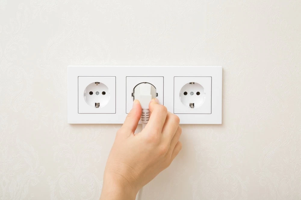 A hand is plugging a white electrical cord into the middle socket of a triple wall outlet on a textured white wall. Two adjacent sockets remain empty.