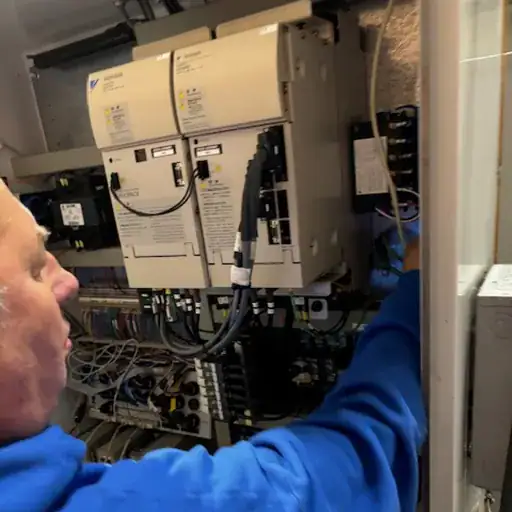 Electrical Panel Repair Taylor County Tx a Person in a Blue Jacket is Working on a Complex Electrical Panel or Server Rack Adjusting or Connecting Wires and Components the Setup Includes Various Cables Connectors and Devices Installed on a Wall
