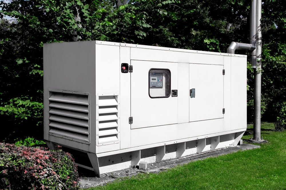A large white outdoor generator sits on a platform surrounded by greenery. It has air vents on the side and a control panel visible through a small window. A metal exhaust pipe extends upward from the unit.