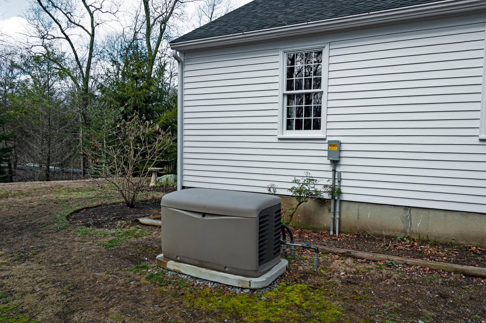 A gray backup generator is installed beside a white house with siding. There's a window above the generator and a utility box nearby. The surrounding yard has some plants, bare soil, and moss. Trees are visible in the background.