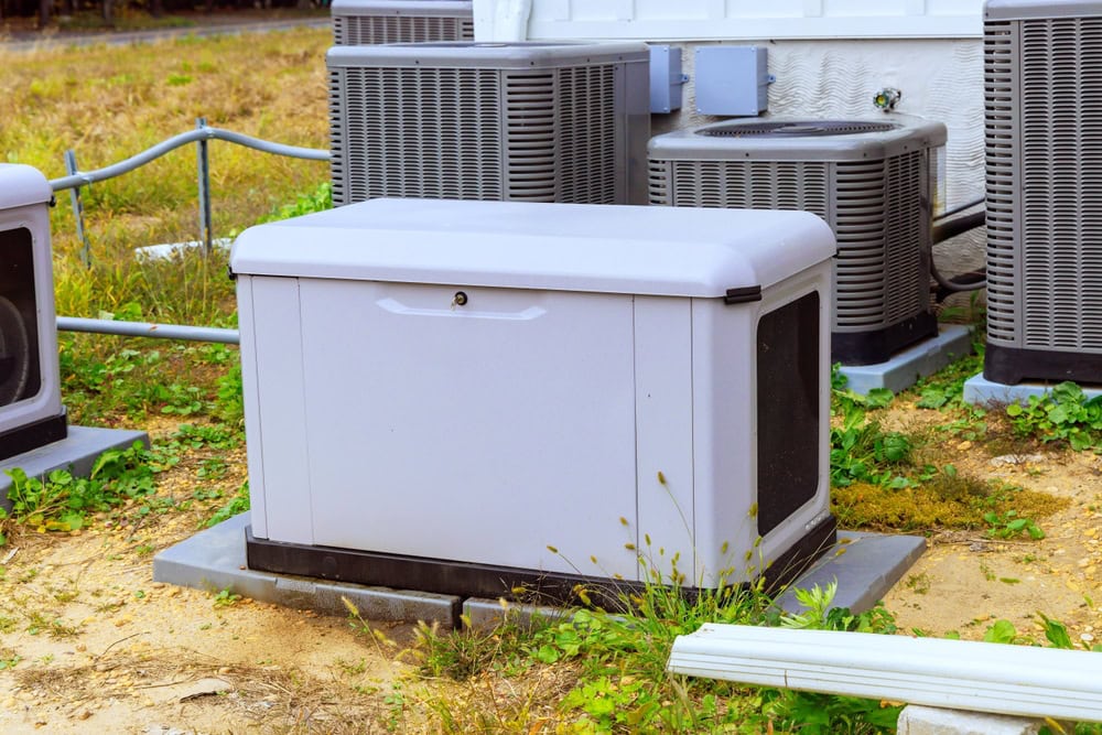 A generator installation is neatly positioned on a concrete slab outdoors, amid several air conditioning units. The area is dotted with patches of grass and dirt, while a white fence peeks through the background.