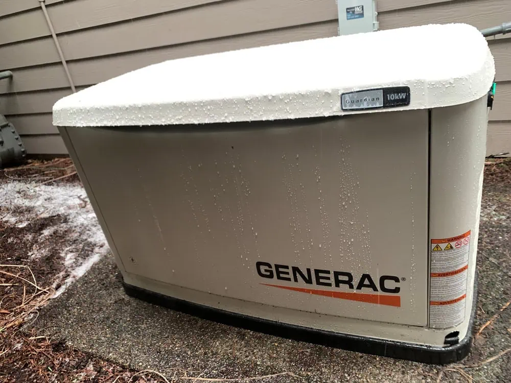 A Generac generator installation is visible outside a building, dusted lightly with snow. The beige unit is marked "Guardian 10kW" and "Generac" in black with an orange line underneath. A wooden siding serves as the backdrop.