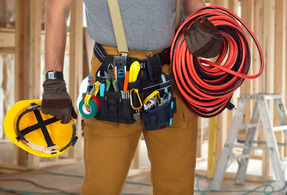 An electrical contractor strides confidently, carrying a yellow hard hat and a coil of red electrical cables. Dressed in protective gloves and khaki pants, a tool belt packed with tools hangs at their waist. Wooden framing and a ladder provide the backdrop to their skilled work.
