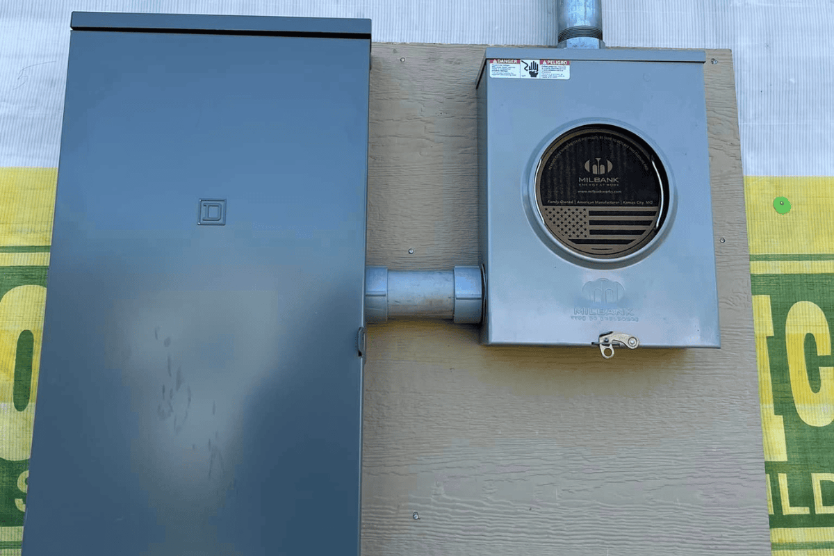 5 a Metal Electrical Box and Meter with a Milbank Logo Are Mounted on a Beige Wall Expertly Installed by Electricians the Meters Transparent Section Reveals Internal Components While Pipes Connect Both Boxes a Yellow and Green Sign is Partially Visible in the Background