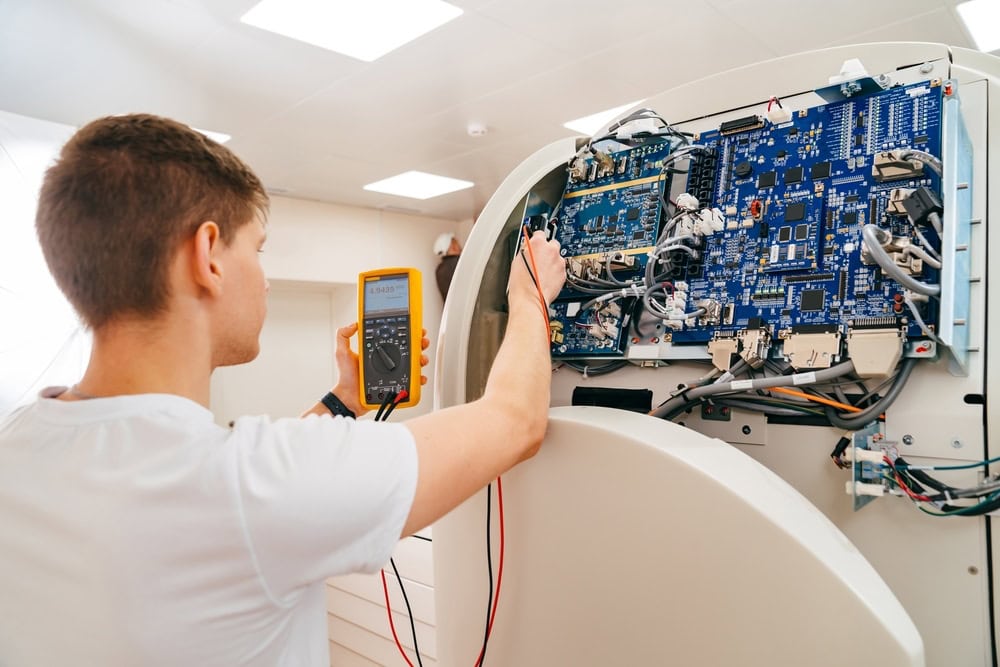 A technician, like electricians in Taylor County, TX, uses a digital multimeter to test or repair a large circuit board inside an open electronic device, surrounded by cables and components in a bright, modern workshop.