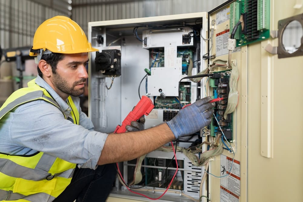 An electrician in TX wearing a yellow hard hat, gloves, and safety vest tests wires with a multimeter on an open electrical panel—showcasing the expertise of electricians Taylor County relies on in industrial settings.