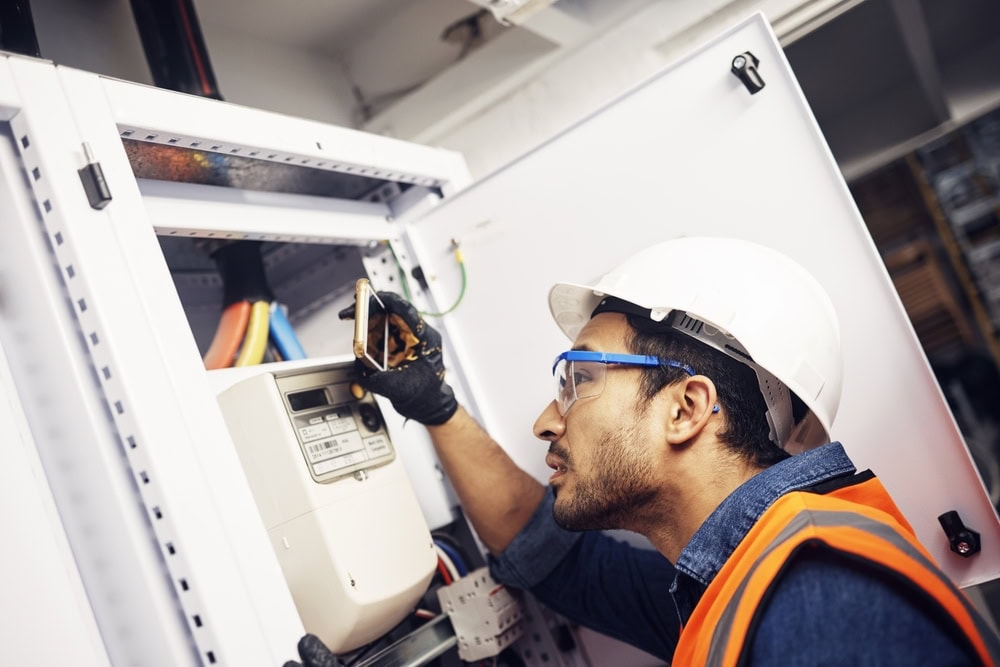 A worker in a hard hat, safety glasses, and an orange vest inspects an open electrical panel with a flashlight, just like skilled electricians in Taylor County, TX, closely examining the wiring and components inside.